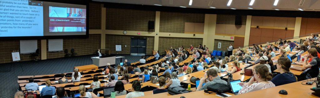 Hastily-cropped photo from the back of the room showing 12 rows of stadium seating with a speaker in the front of the room in front of one large screen for slides and another for captions.