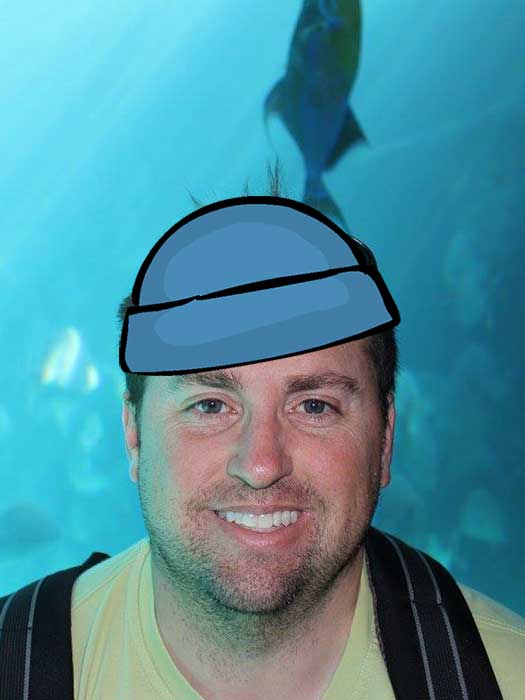 Travis Brown with a cartoon blue beanie in front of a large fish tank, perhaps channeling “The Life Aquatic.”