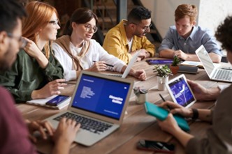 A diverse group of young adults working together on tablets and laptops while sitting at a conference table.