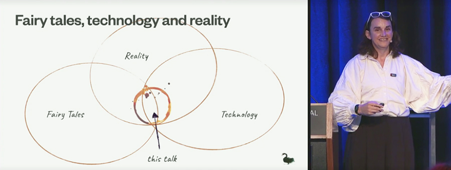 Tea Uglow stands next to a slide showing three intersecting circile labeled Fairy Tales, Reality, and Technology. At their intersection is a circle formed by a coffee cup stain labelled This  Talk. 