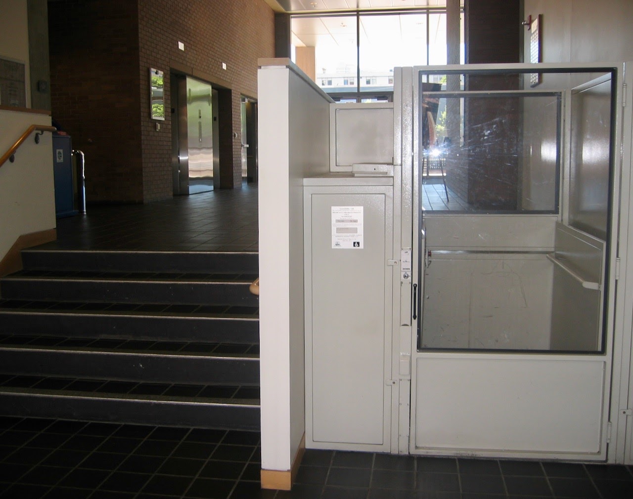 Photo with platform lift on the right and five-step staircase with railing on the left. The stairs and lift are inside an entrance hall with high ceilings and windows.